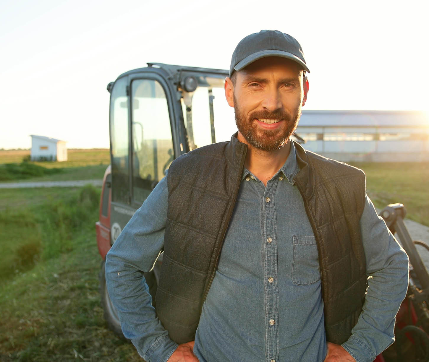 Portrait of young Caucasian handsome happy man farmer standing in field and smiling to camera. Big tractor on background. Cheerful male worker in agricultural farm. Sunlight. Agriculture farming.